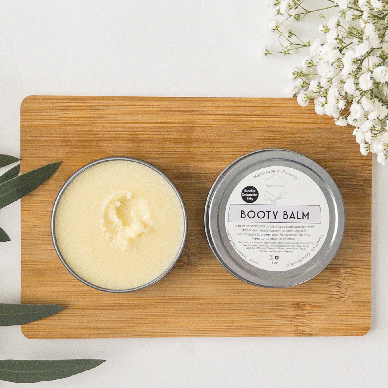Trades of Brit Booty Balm open tin with baby breath and eucalyptus around it.