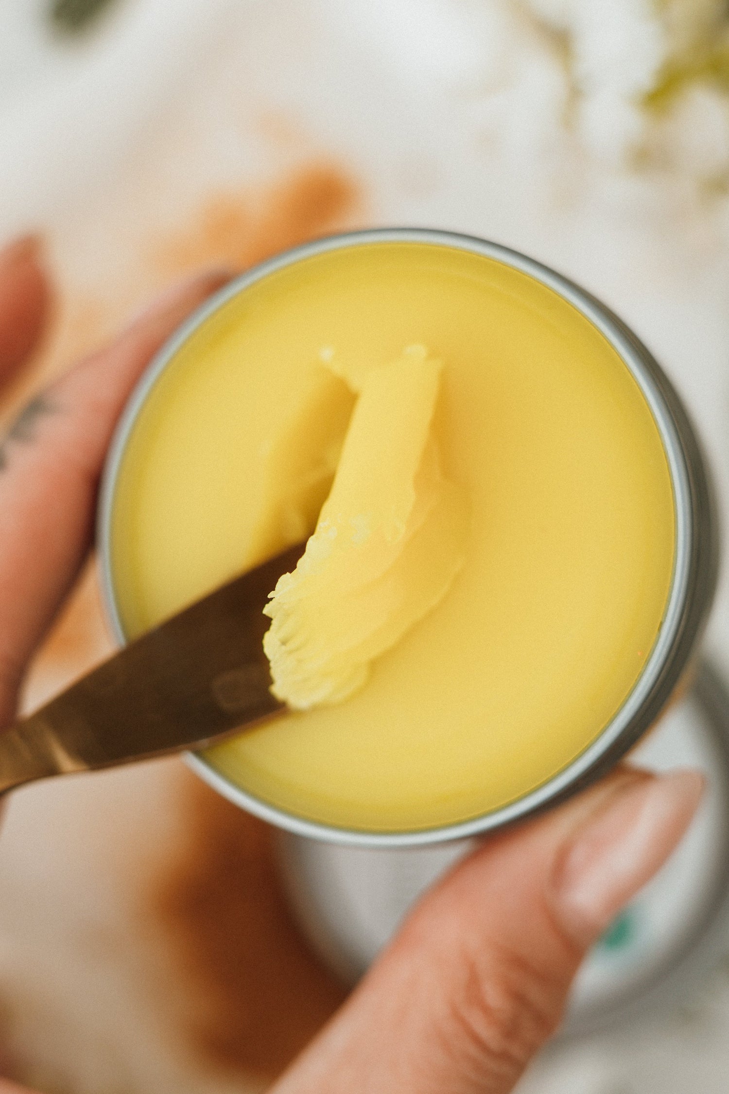 Hand holding a tin contain salve with a metal spatula in the tin. 