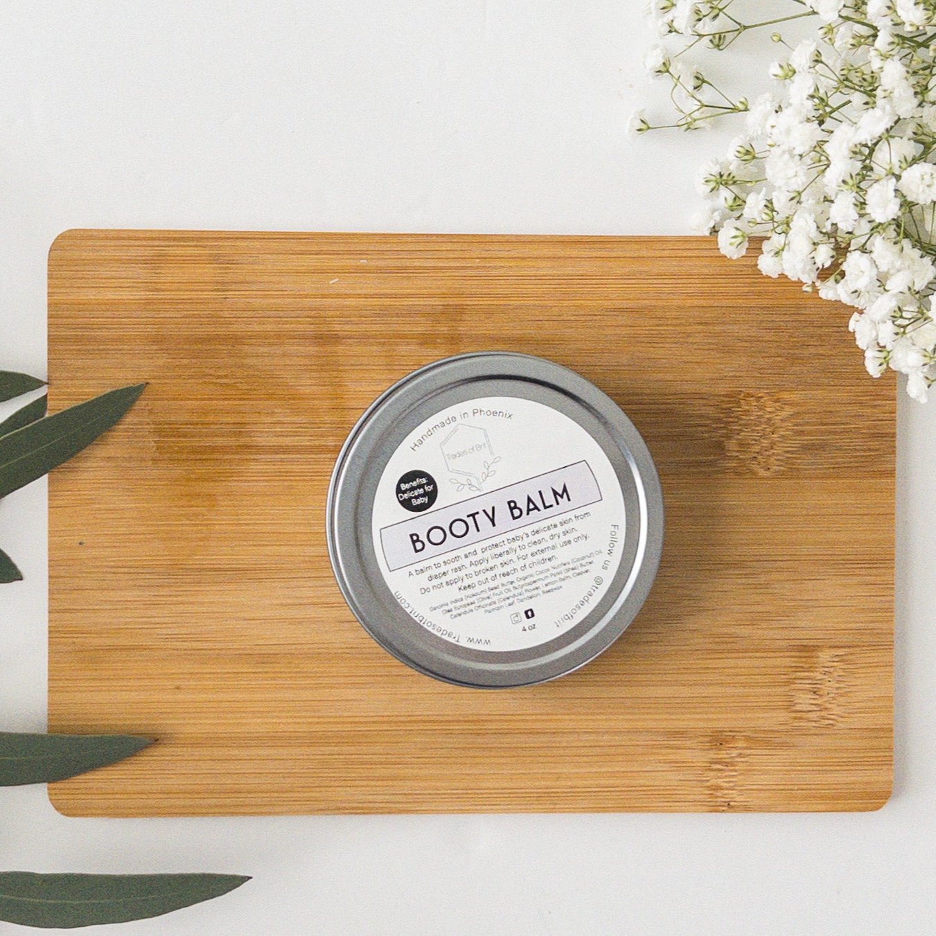 Trades of Brit Booty Balm Tin on a wooden board with baby breath flowers and eucalyptus around it. 