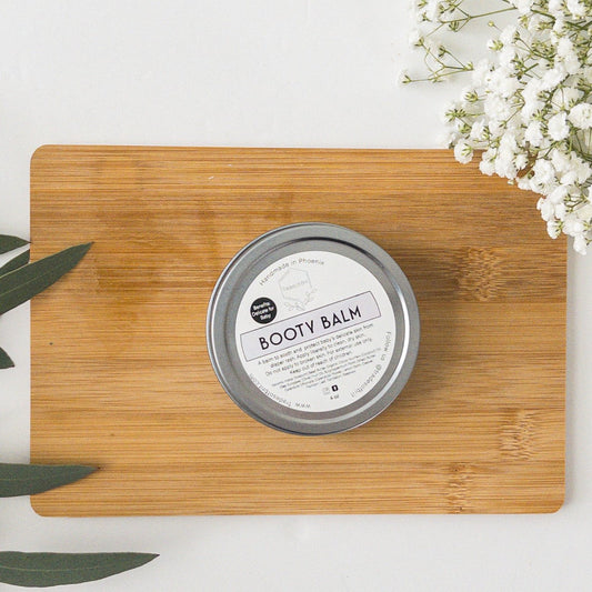 Trades of Brit Booty Balm Tin on a wooden board with baby breath flowers and eucalyptus around it. 