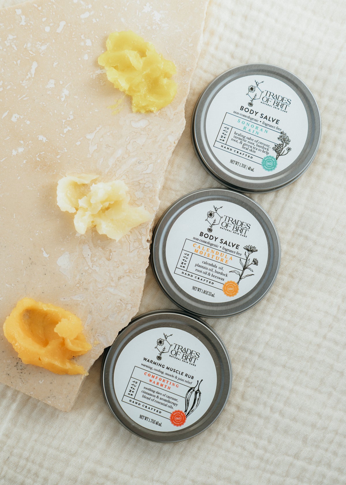 Three small round tins of Trades of Brit body salves arranged on a textured white fabric beside a stone slab with yellow balm pieces. The visible labels show different scents: Sonoran Rain, Calendula and Warming Muscle Rub.